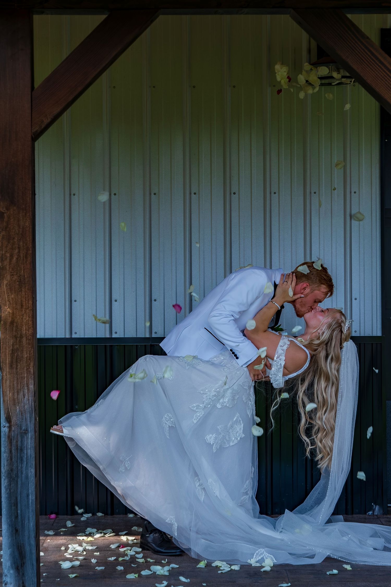 A romantic dip pose between a couple in wedding attire against a rustic wall with scattered flower petals.