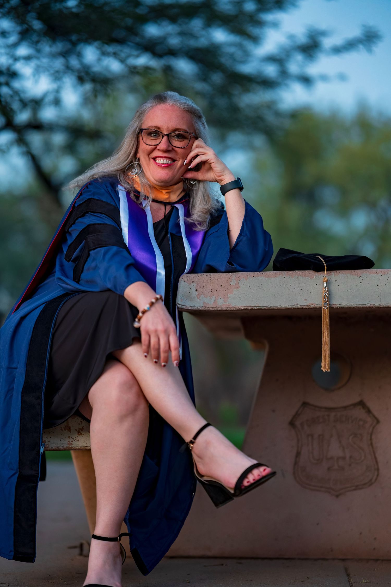 Graduate in blue and purple regalia sits casually on bench during outdoor portrait session.