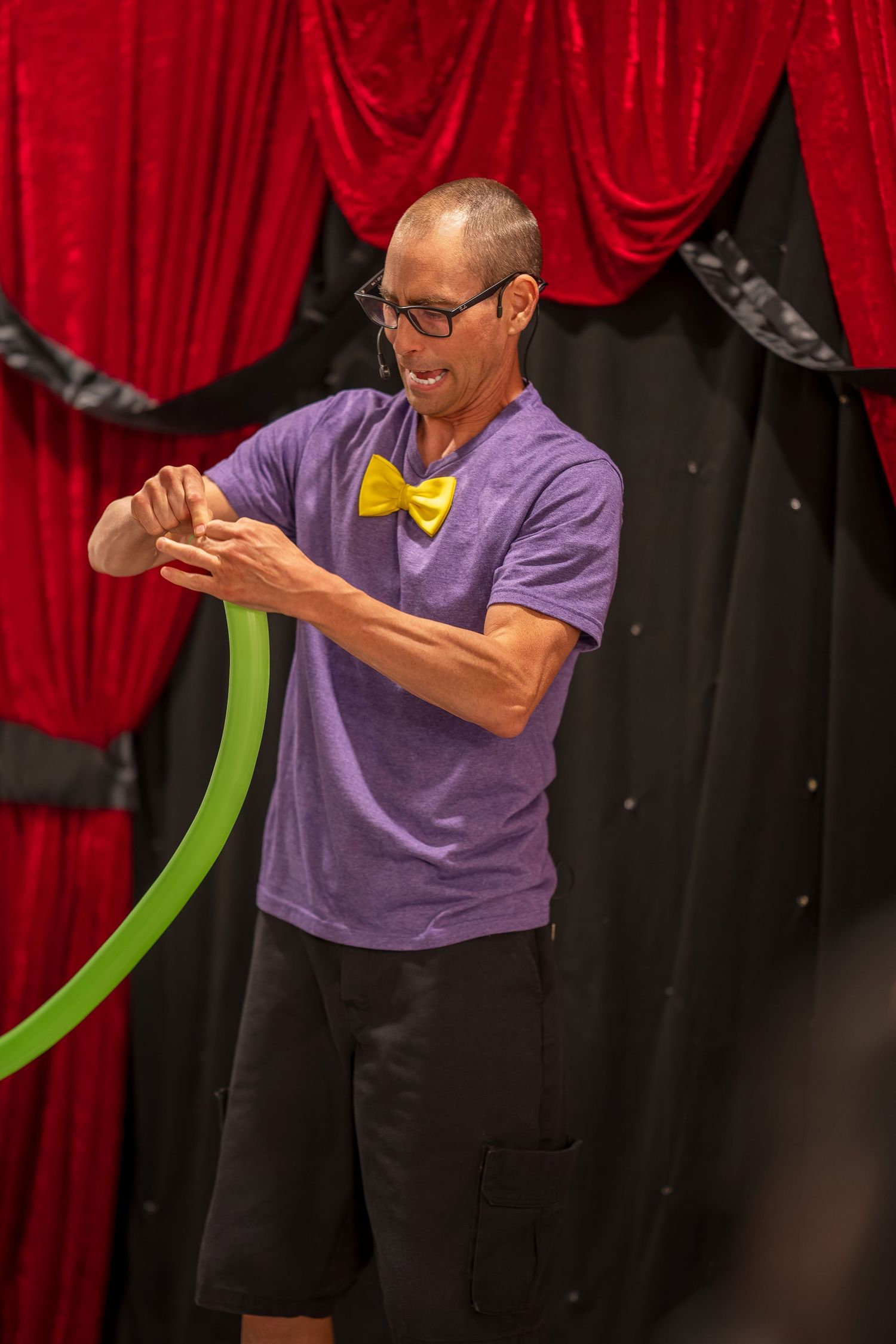 Magician in purple shirt performing with props against red curtain backdrop.