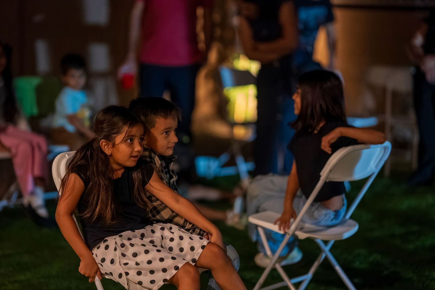 Children sitting on folding chairs at evening outdoor event.