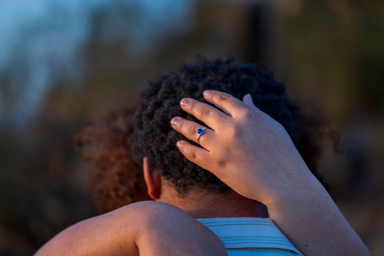 Close-up of hand with blue sapphire ring resting in curly hair during romantic outdoor engagement session.