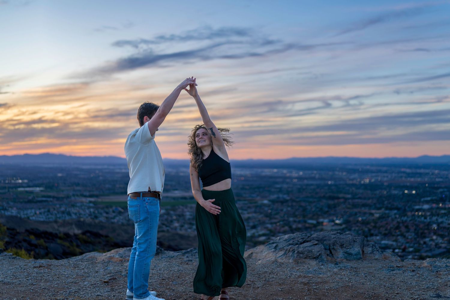 A couple dances joyfully on a hilltop at twilight with a breathtaking cityscape below.