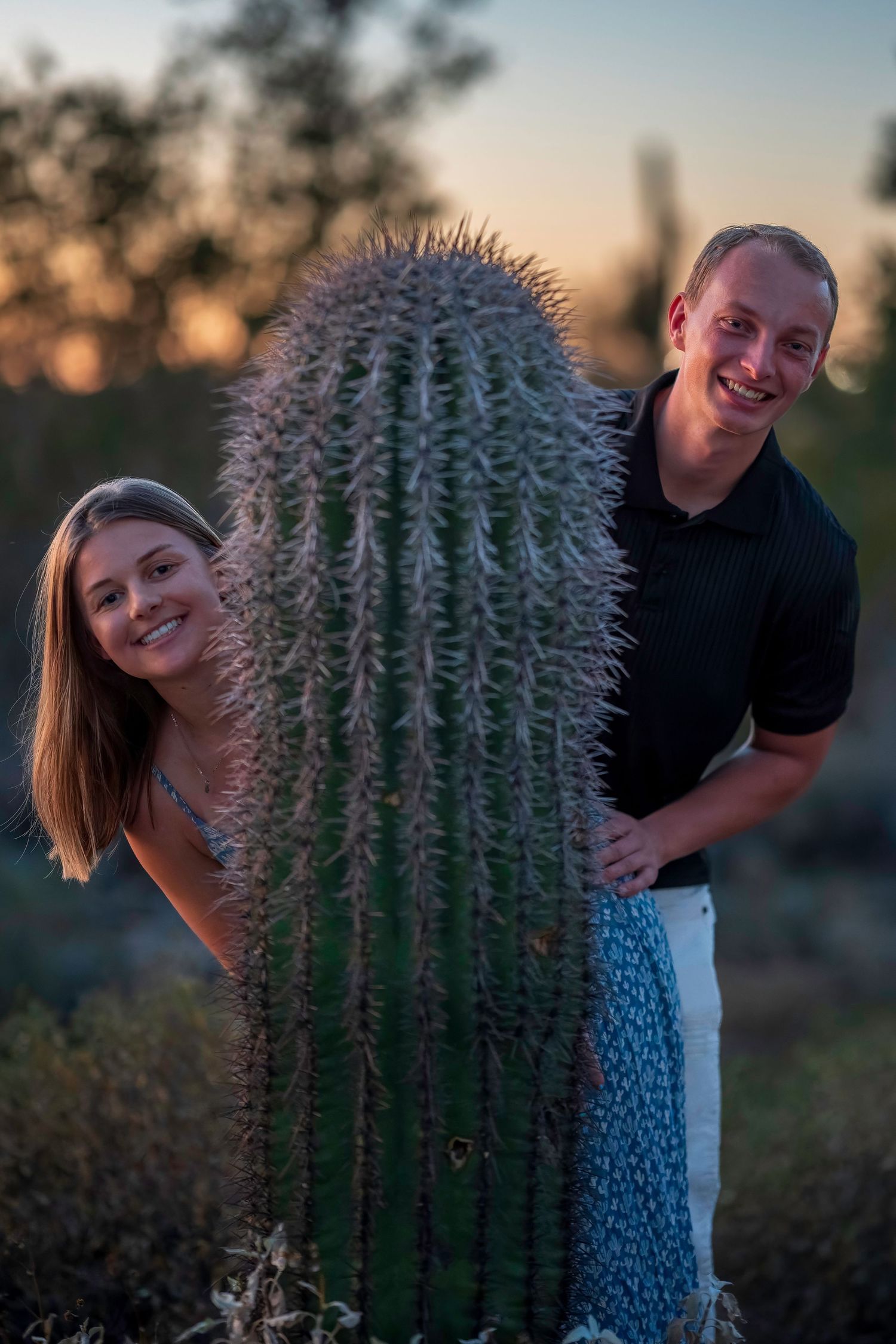 Happy couple smiling behind large saguaro cactus at desert sunset.