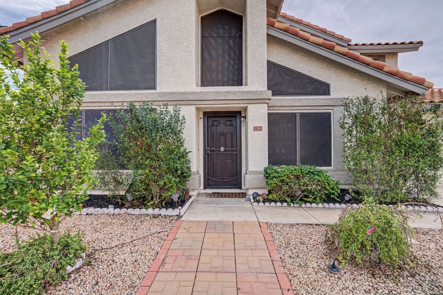 Charming home exterior with brick pathway, desert landscaping, screened windows, and a welcoming dark wood front door.