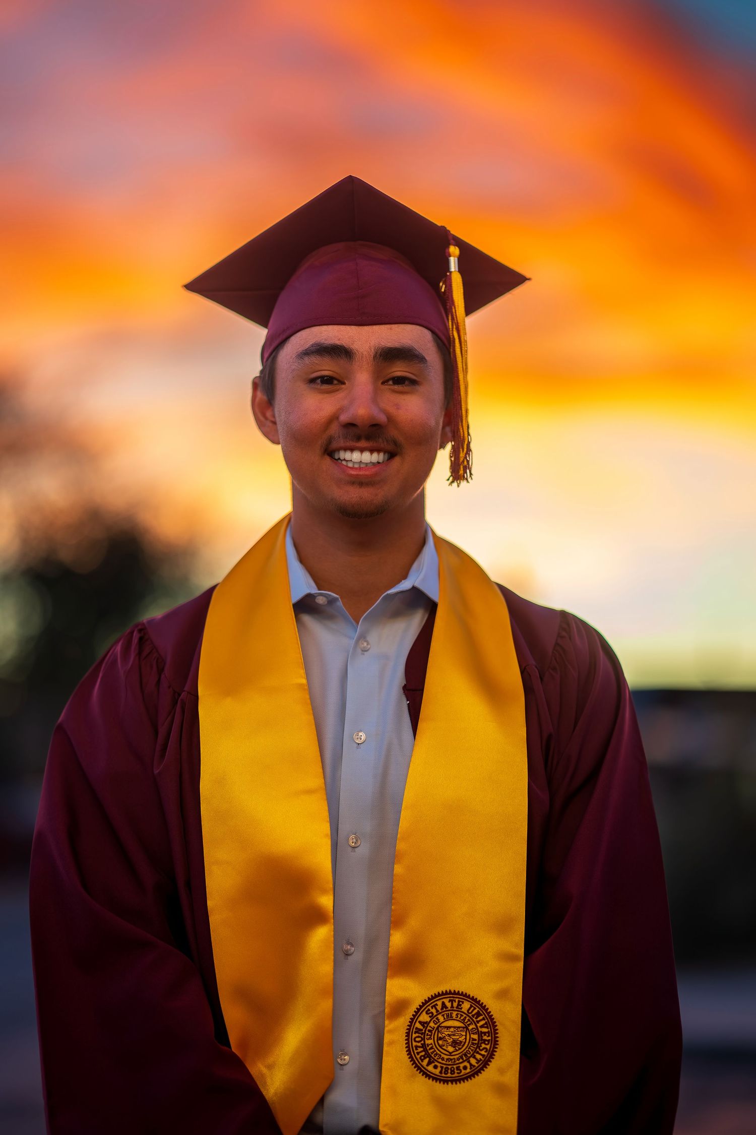Graduation portrait with vibrant orange sunset backdrop and maroon academic regalia.