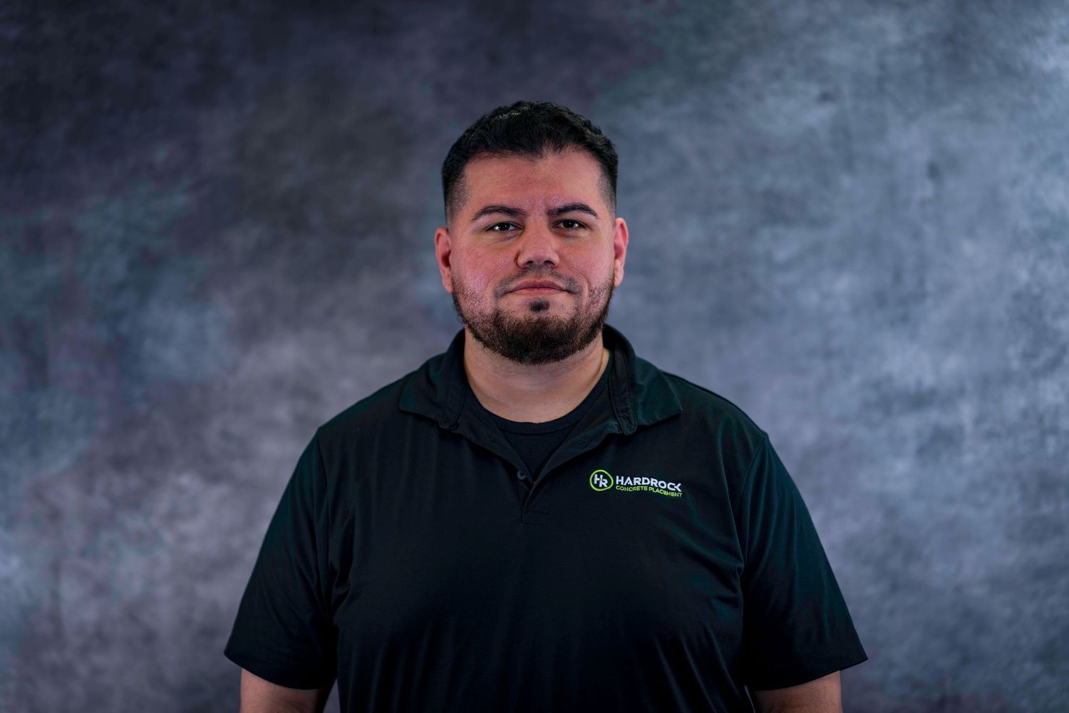 Team member wearing black polo with company branding against dark studio background.
