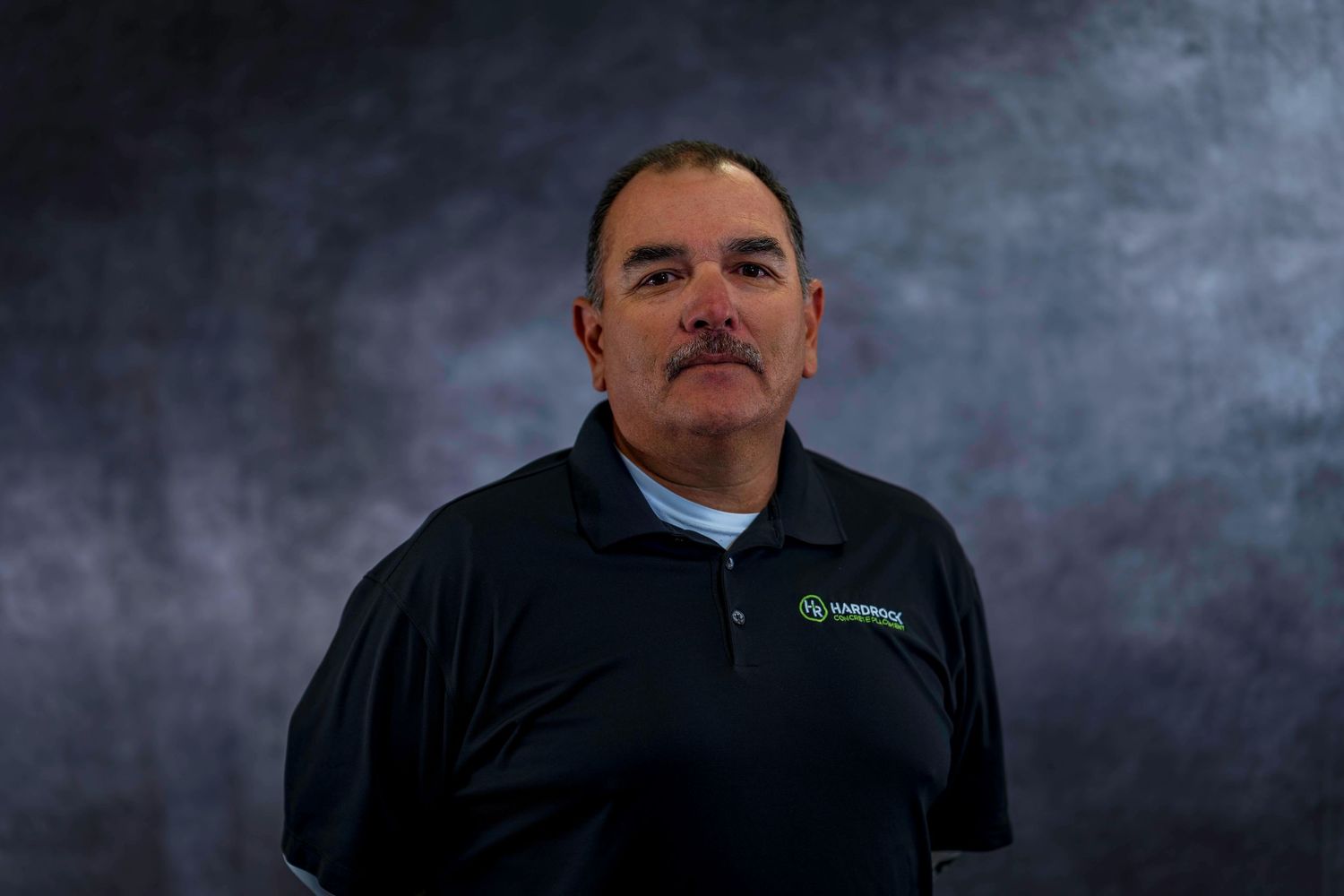 Corporate headshot of person in dark polo with company logo against smoky background.