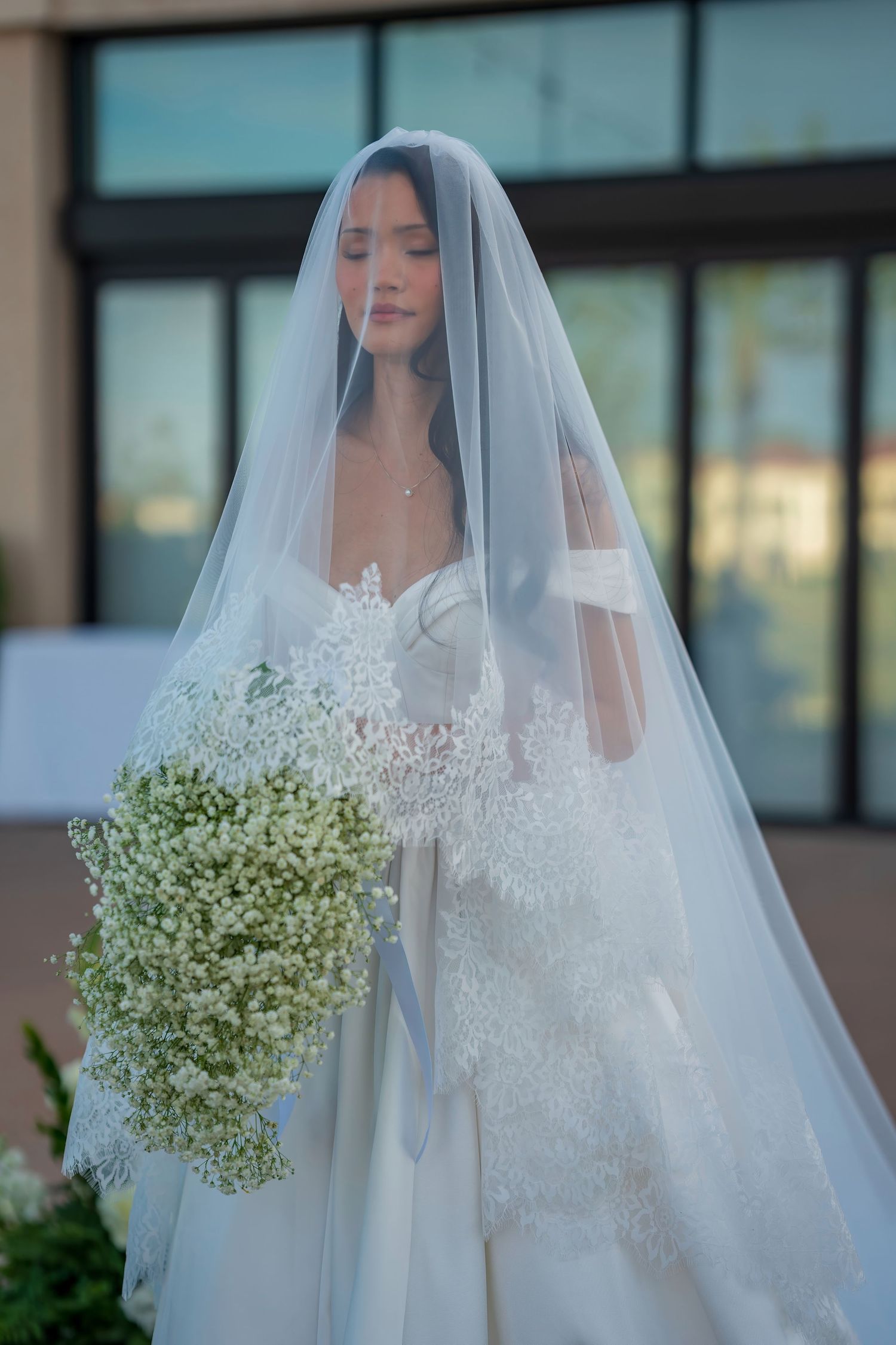 A close-up portrait of a bride wearing a detailed lace gown and sheer veil holding a cascading bouquet.