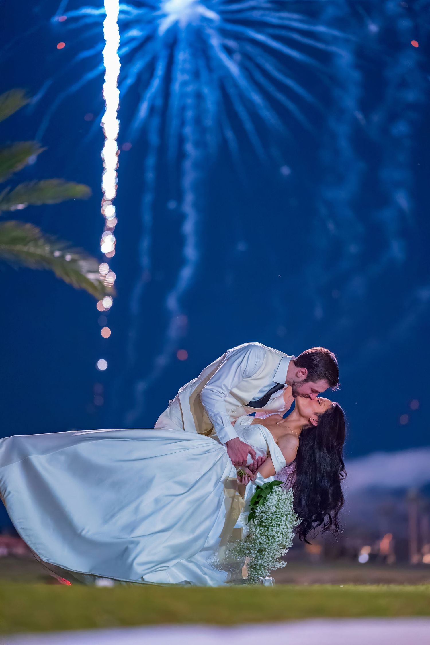 Couple in wedding attire performs romantic dip pose against backdrop of blue fireworks.