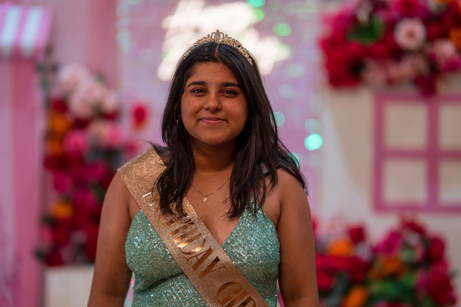 Person in a sparkly turquoise dress posing against a pink floral backdrop at a celebration.