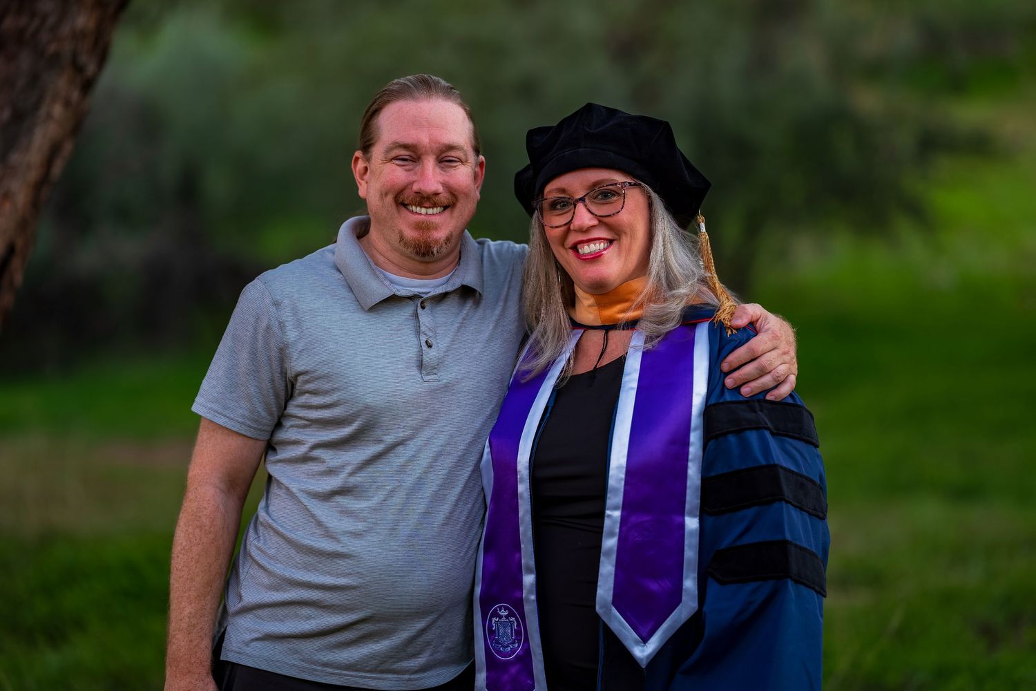 A graduation celebration photo showing a graduate in blue and purple academic regalia with a companion outdoors.