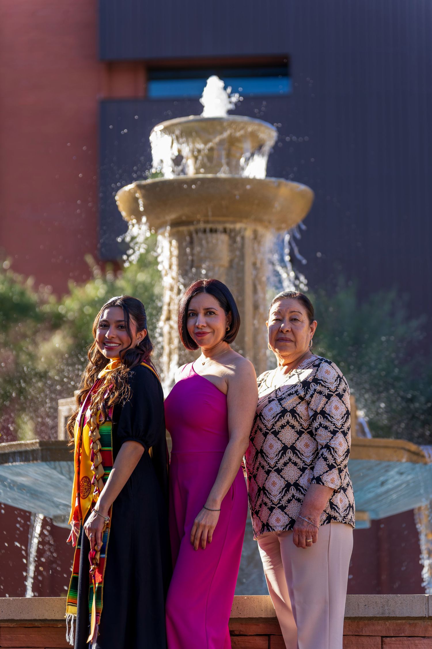 Three people smiling and laughing together near a decorative fountain outside a brick building.