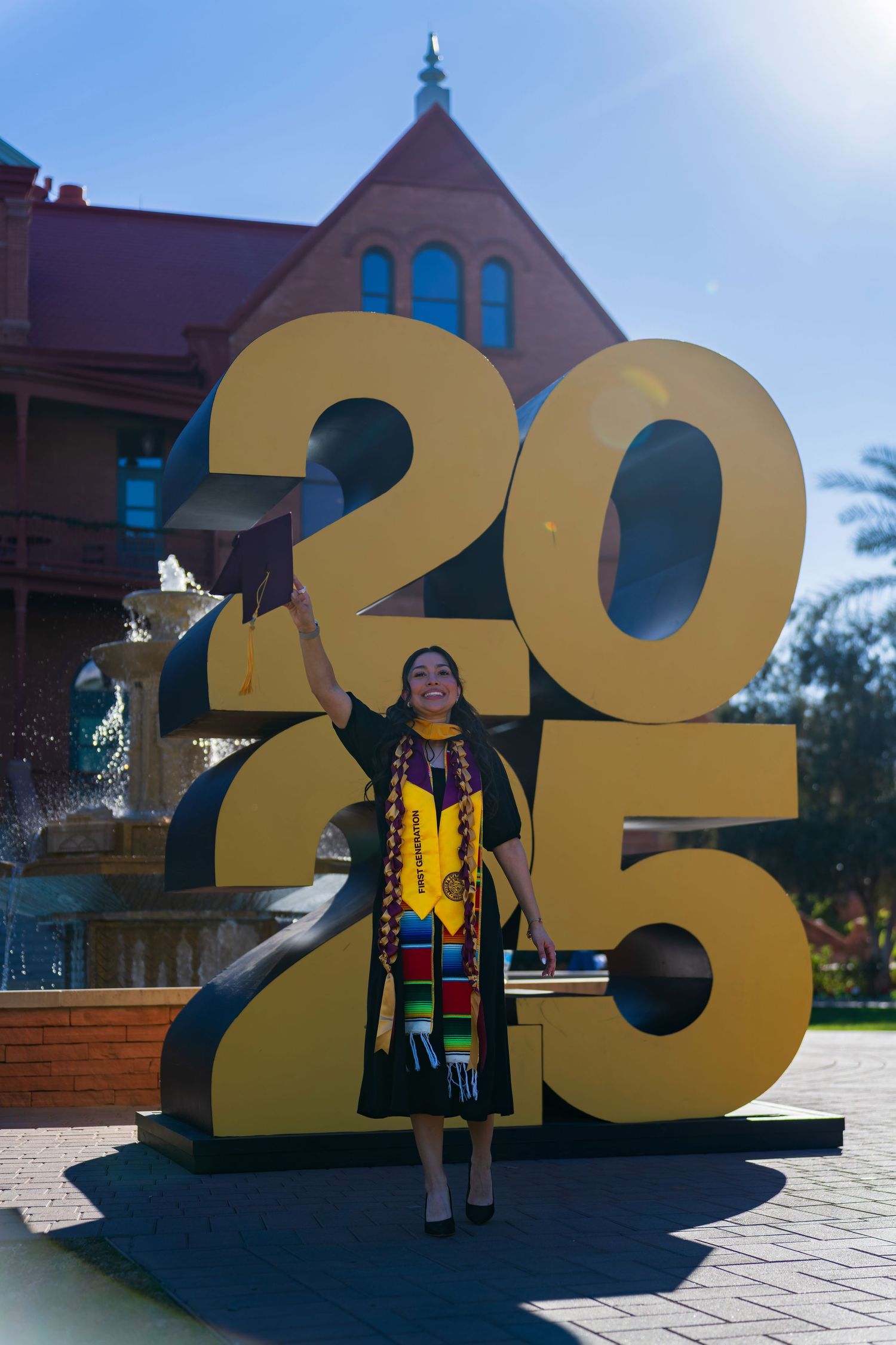 Graduate wearing traditional stole and cap stands by gold 2023 display on sunny day.