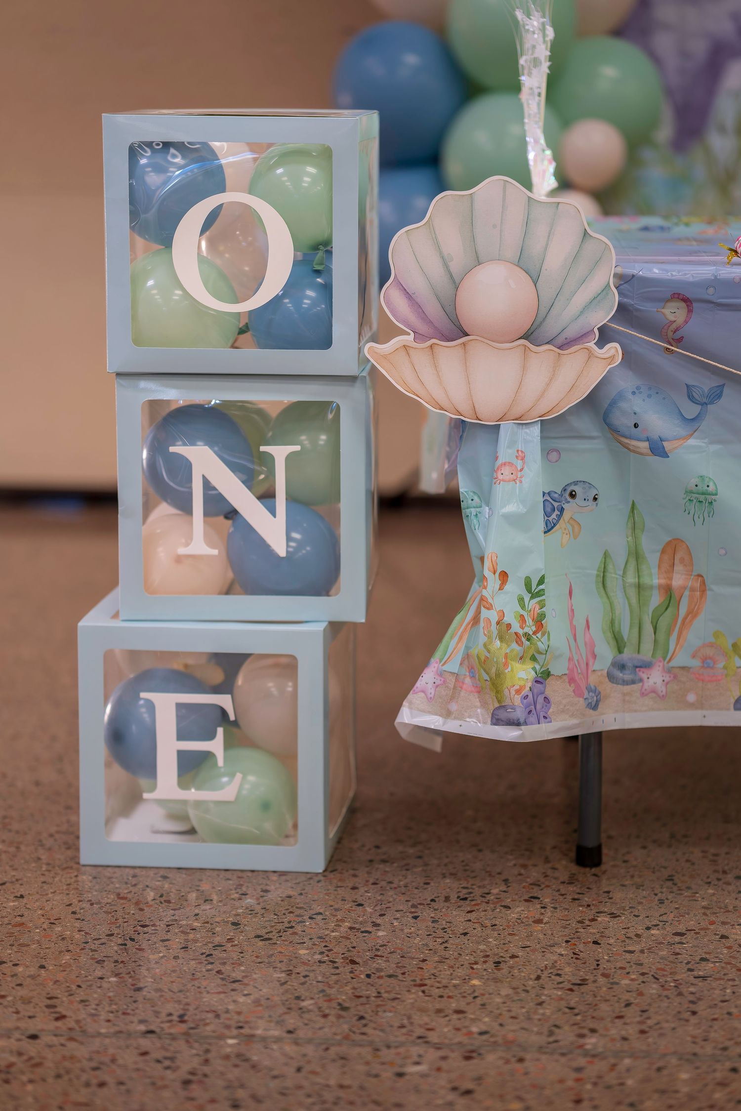 Transparent letter blocks spelling ONE filled with blue and green balloons beside a mermaid-themed first birthday party decoration.