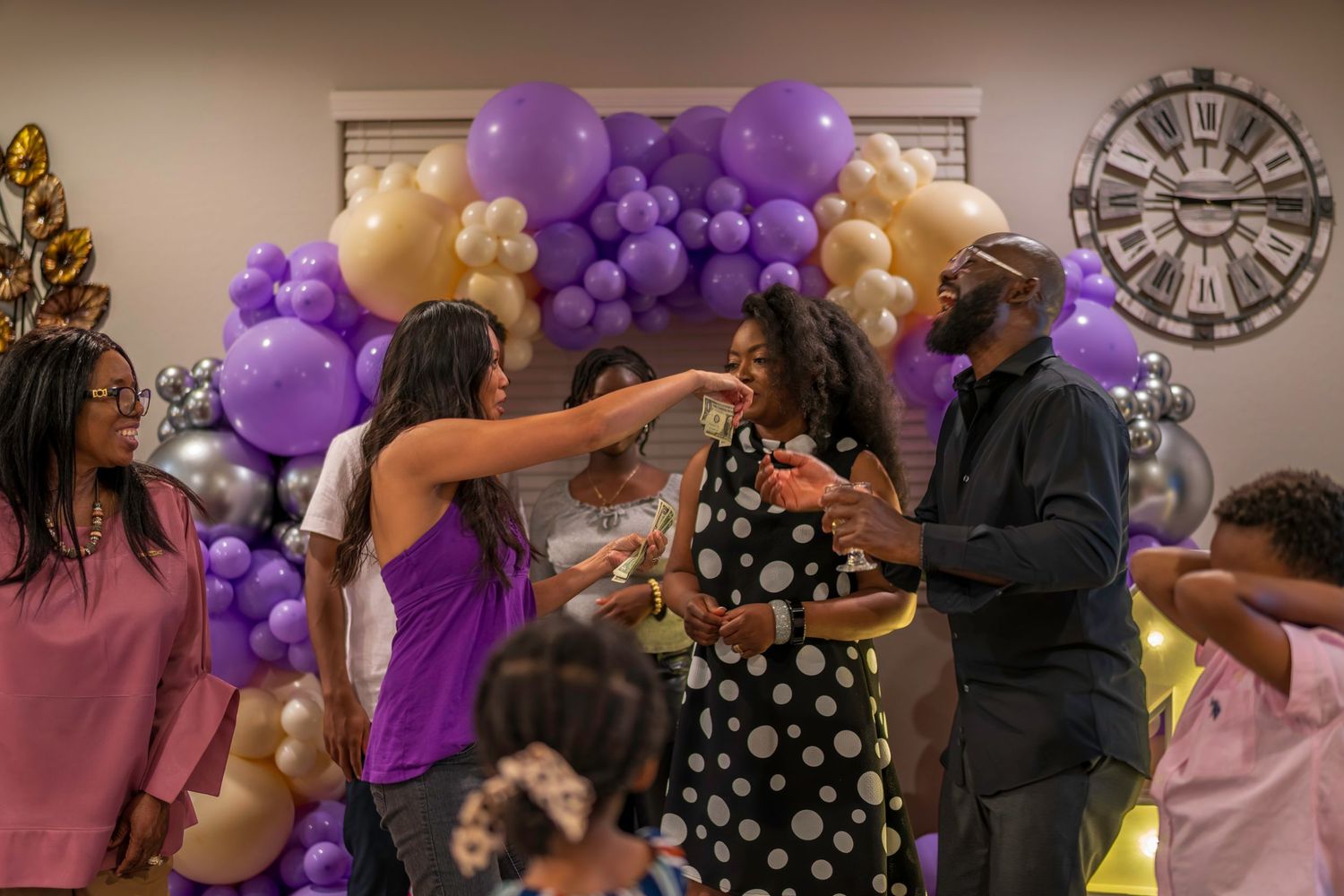 People celebrating at a party with purple, gold, and silver balloon arch backdrop in decorated indoor venue.