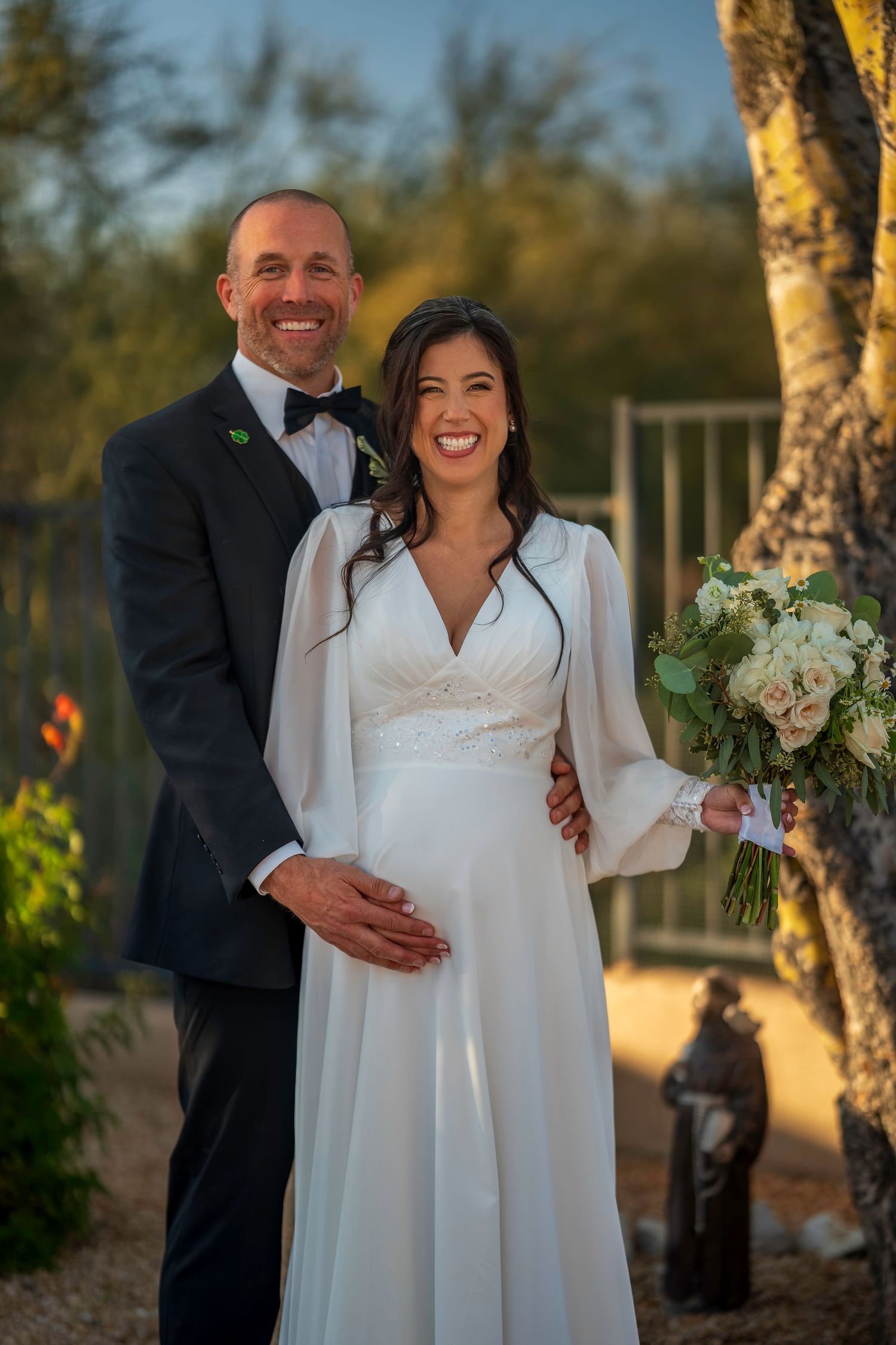 Smiling bride and groom pose together at golden hour, bride holding white rose bouquet outdoors.