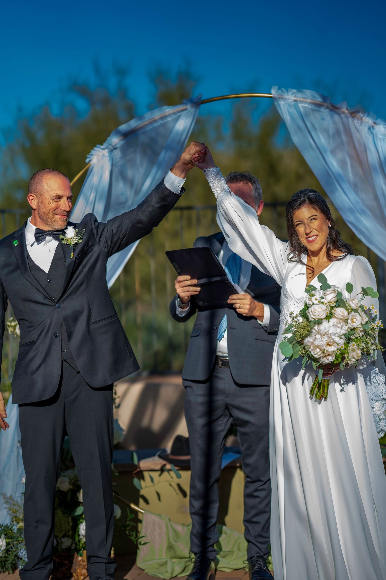 Bride and groom raise joined hands triumphantly at outdoor wedding ceremony beneath blue draped arch.