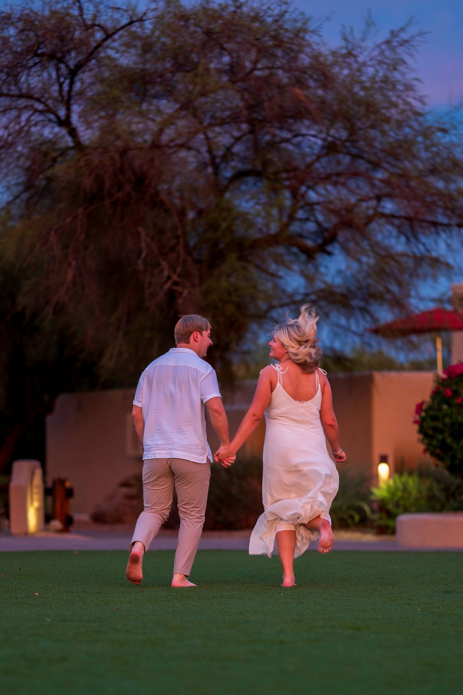 Couple holding hands walking across green lawn at dusk in white wedding attire.