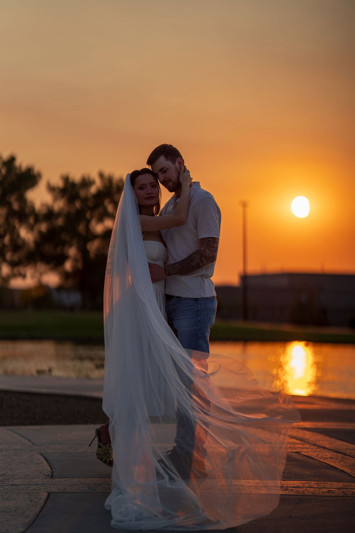 A bride and groom embracing at sunset near a reflective pool, her veil flowing in the wind.