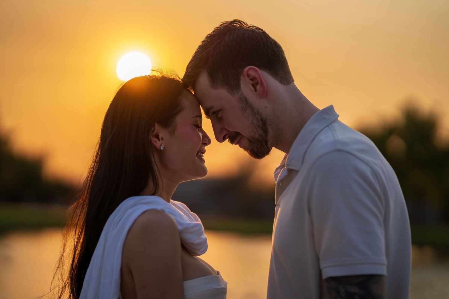 A couple leans forehead to forehead smiling tenderly against a vibrant orange sunset.