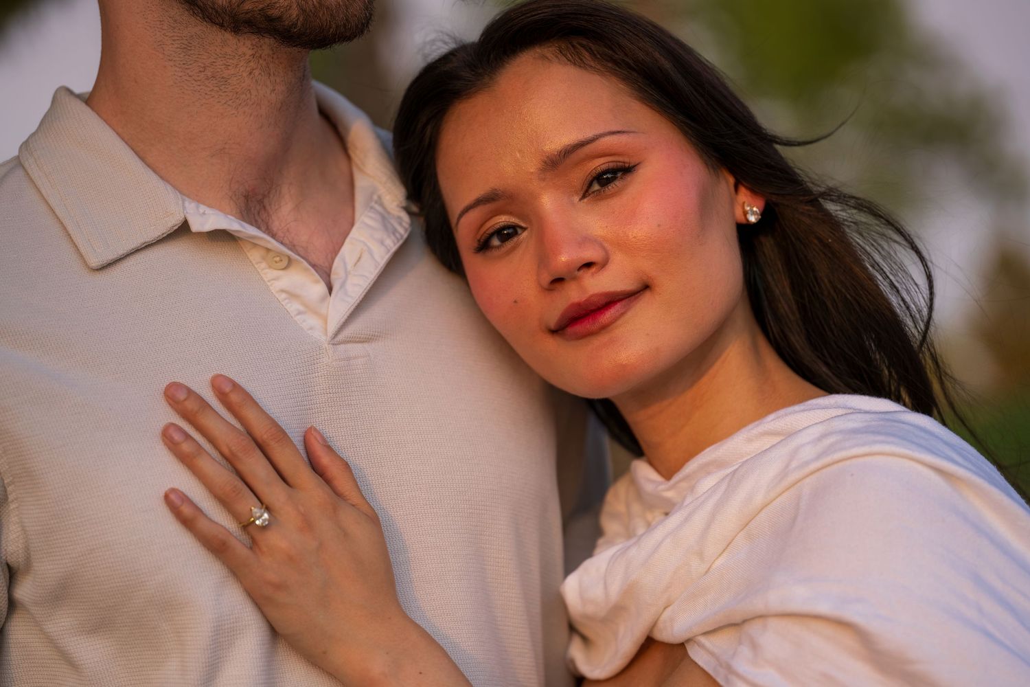 A woman with an engagement ring rests her head on her partners chest outdoors.