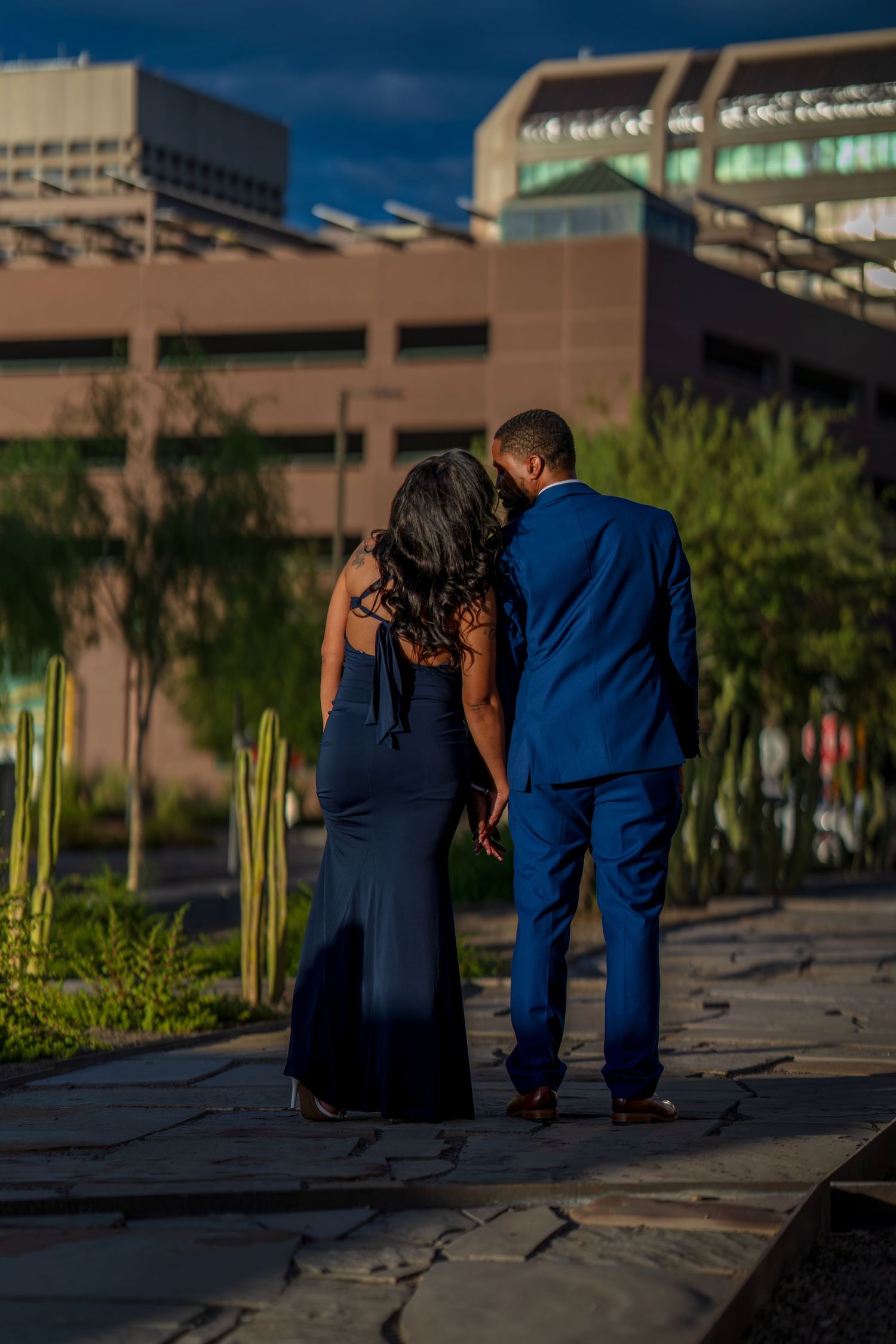Couple in matching navy blue formal attire walking hand-in-hand along outdoor path at sunset.