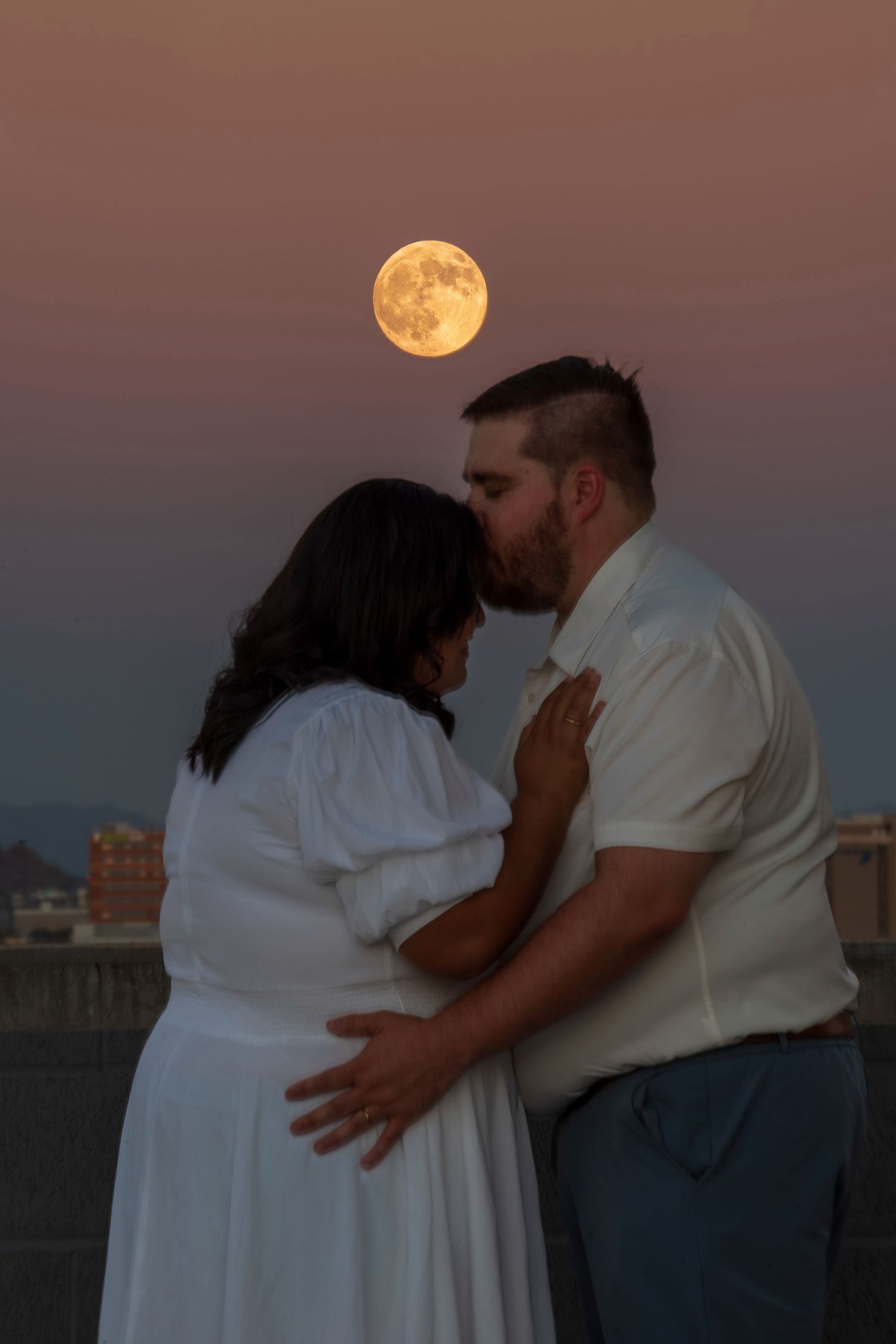 Couple sharing a romantic kiss under a full moon at dusk, woman in white dress with visible baby bump.