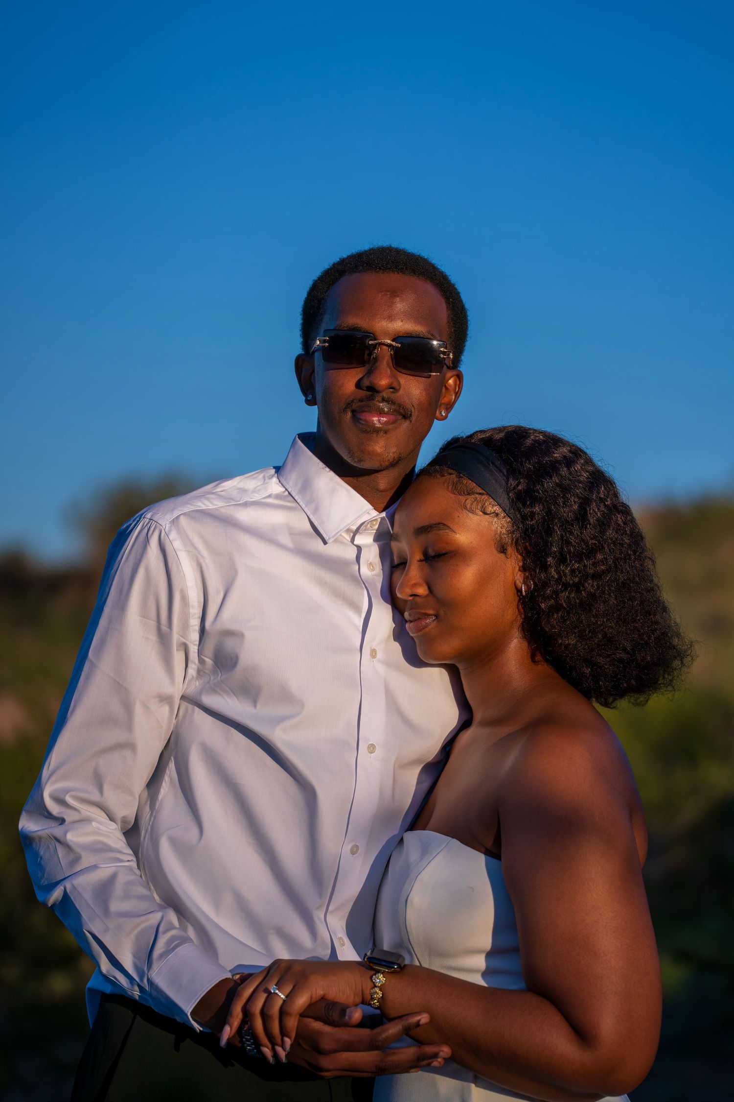 A couple embraces tenderly outdoors against a vivid blue sky, man in white shirt with sunglasses.