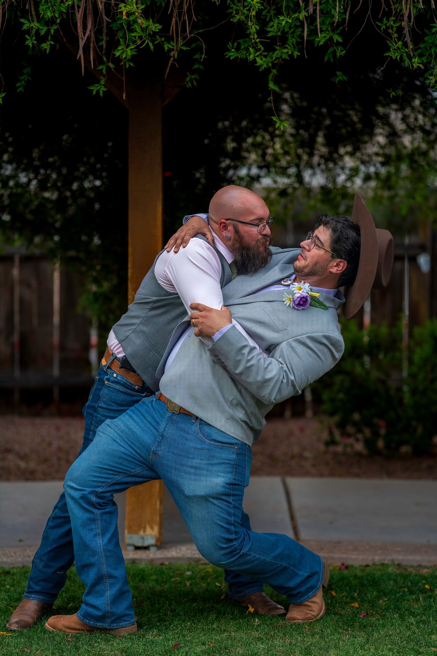 Two men in wedding attire share a joyful embrace outdoors, one wearing a boutonniere and cowboy hat.