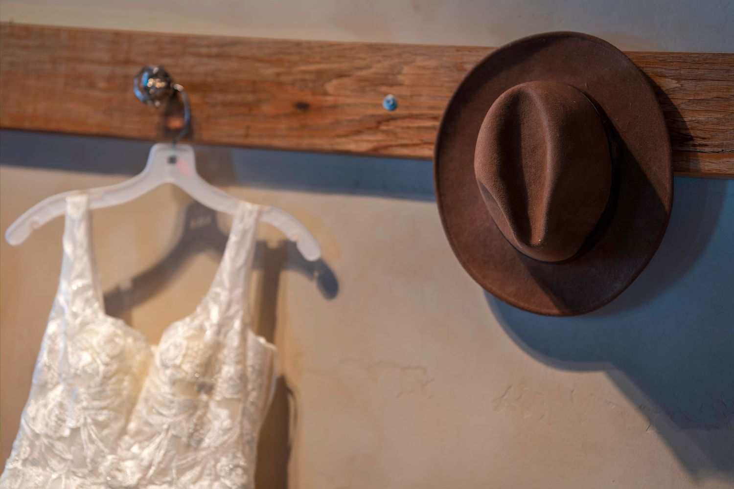 A brown fedora hat and white lace dress on hanger displayed on a rustic wooden wall-mounted coat rack.