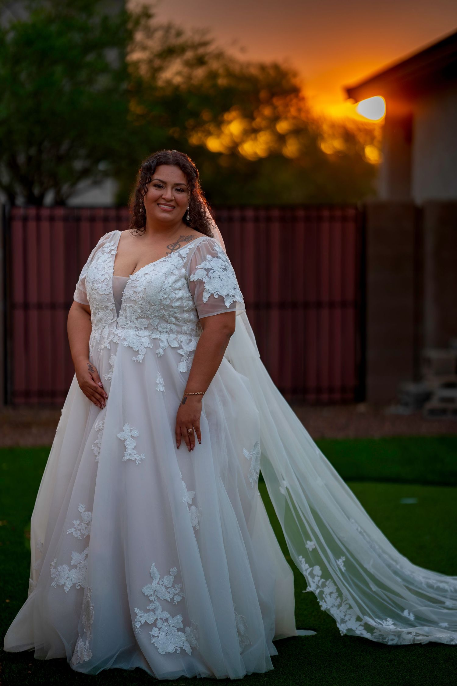 Plus-size bride in white lace A-line wedding gown with floral appliques poses outdoors at golden sunset.