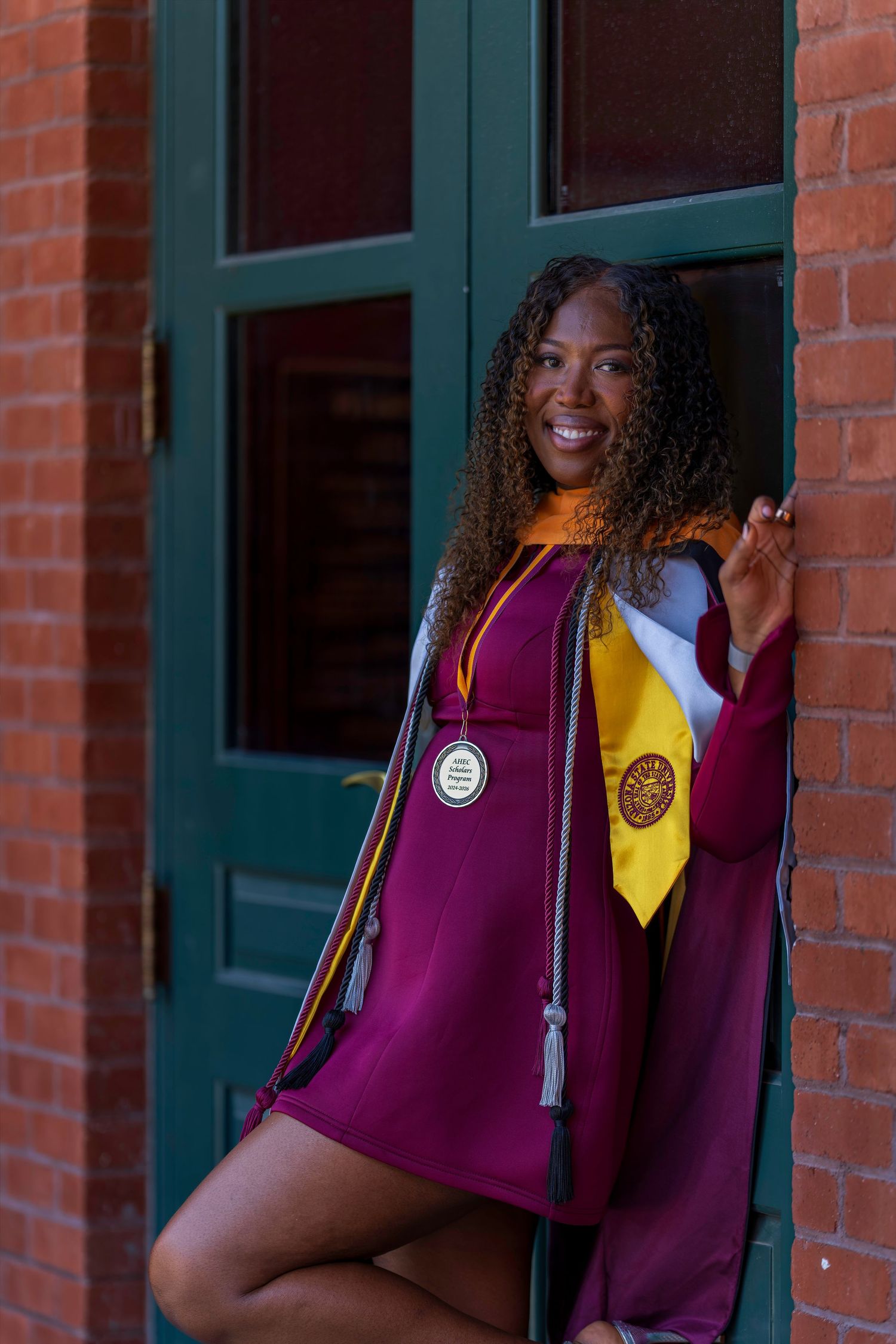Smiling graduate in maroon cap and gown with gold stole and medal poses against brick wall with green door.