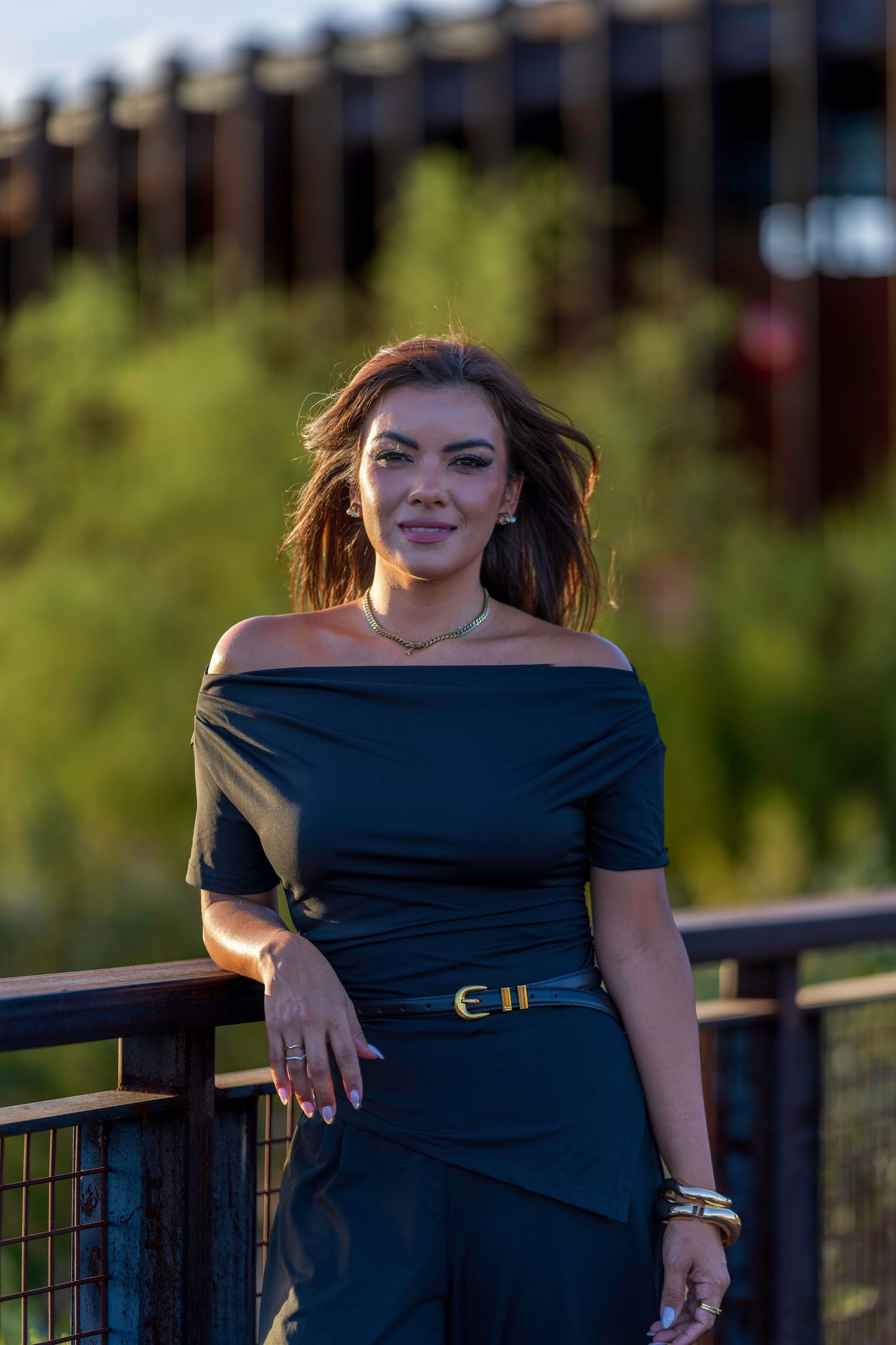 Woman in black off-shoulder top and belt poses confidently on outdoor bridge with lush green background.