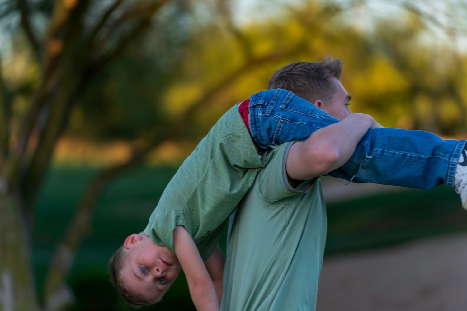 Person in green shirt playfully lifts child upside down outdoors.