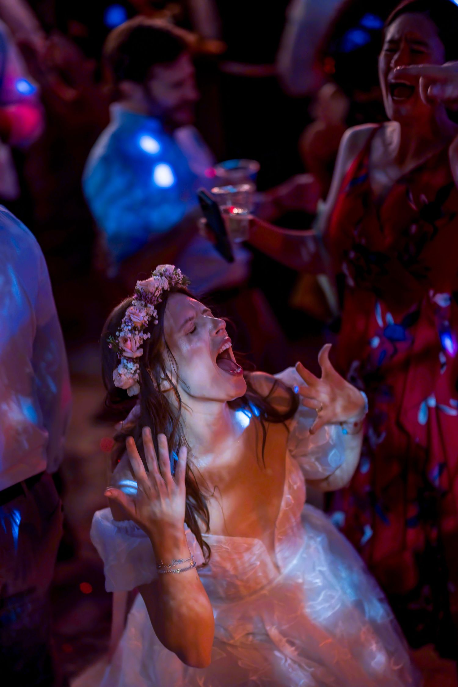 Bride in floral crown dancing joyfully at wedding reception with colorful lights and guests celebrating.