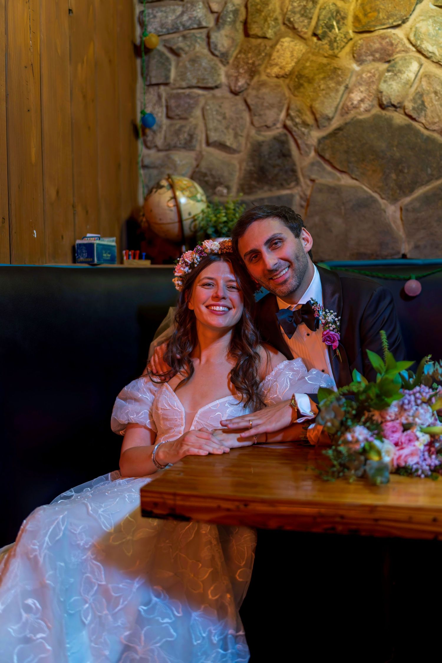 Smiling bride in floral crown and groom in suit sitting together at wooden table with bouquet against stone wall.