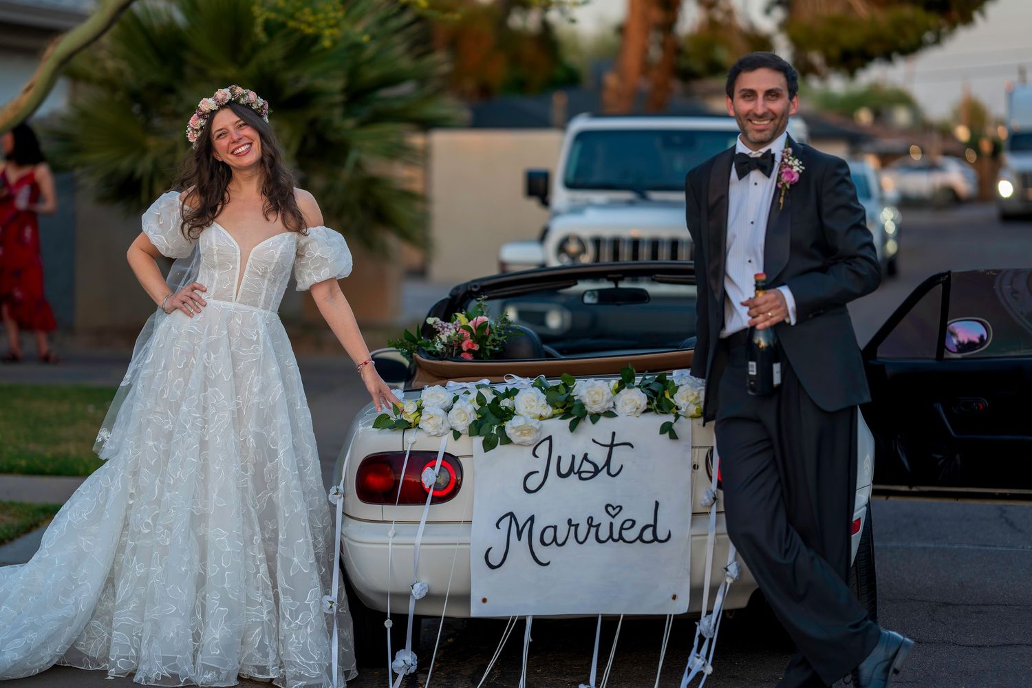 Newlywed couple smiling beside a Just Married decorated white car in wedding attire.