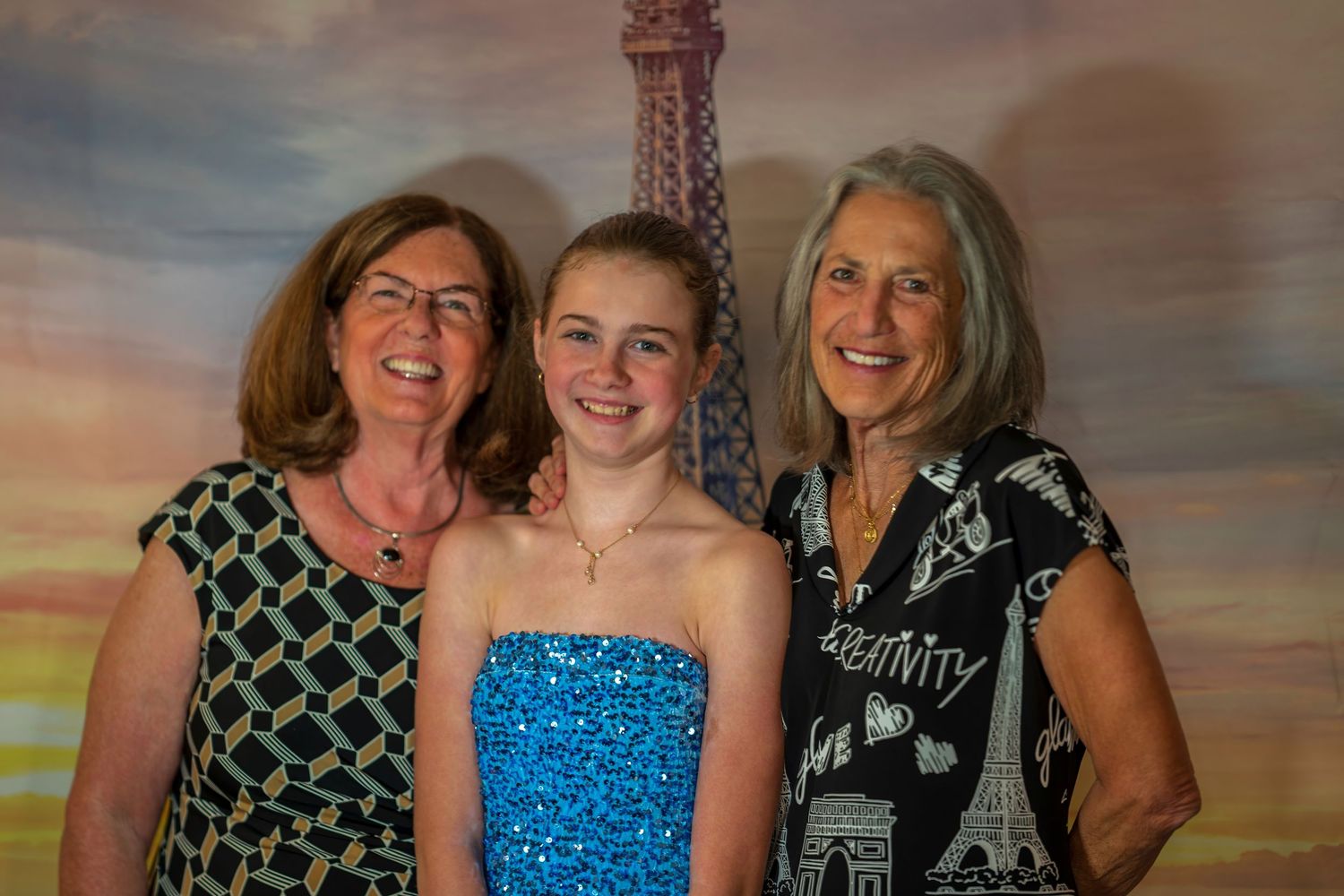 Three generations of women smile together in front of an Eiffel Tower backdrop at a formal event.