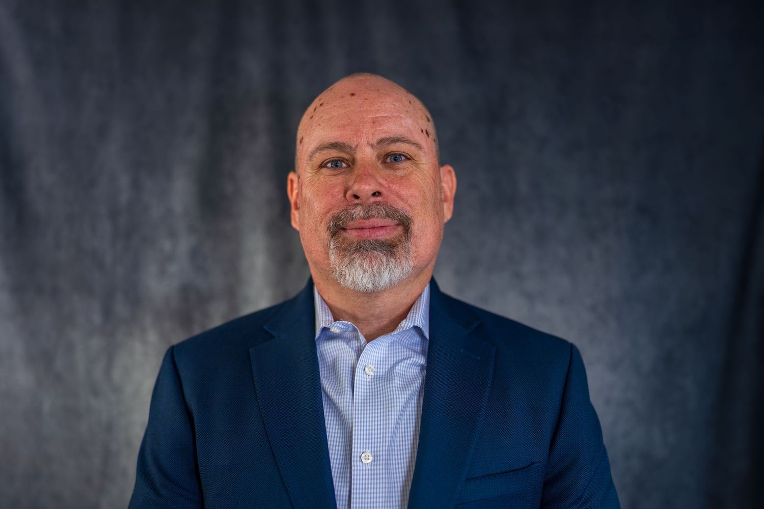Professional headshot against textured gray background wearing navy suit and white shirt.