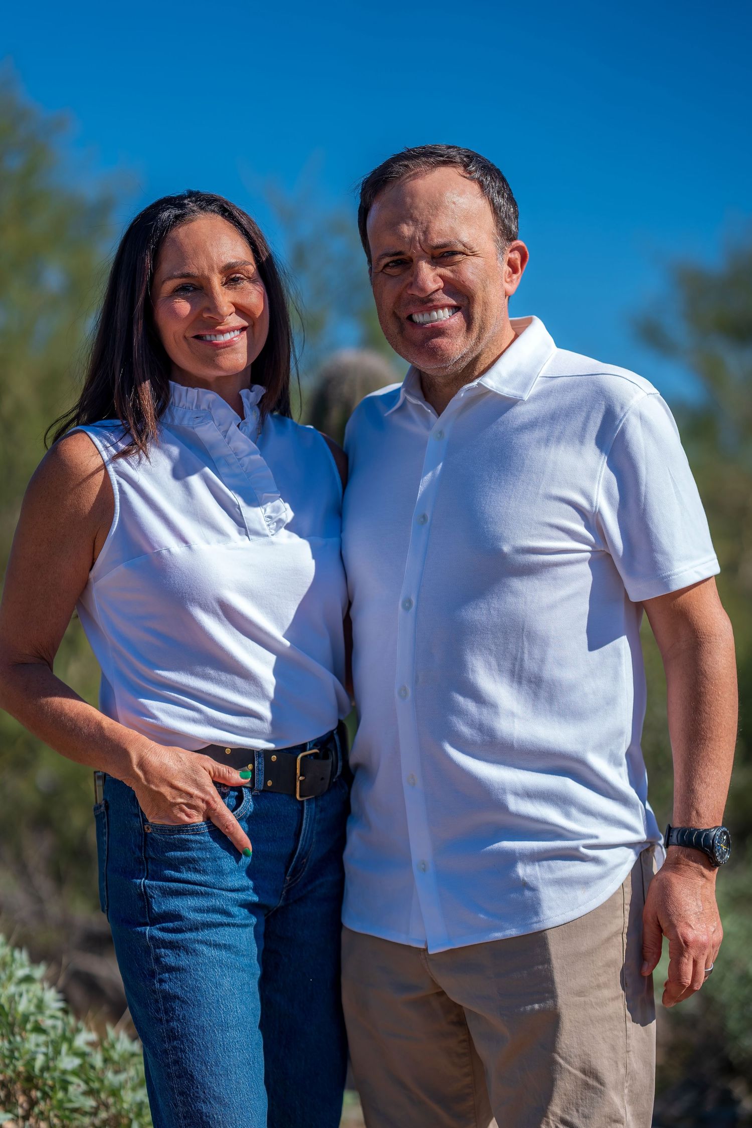 Cheerful couple in coordinated white shirts pose together in a sunny desert setting.