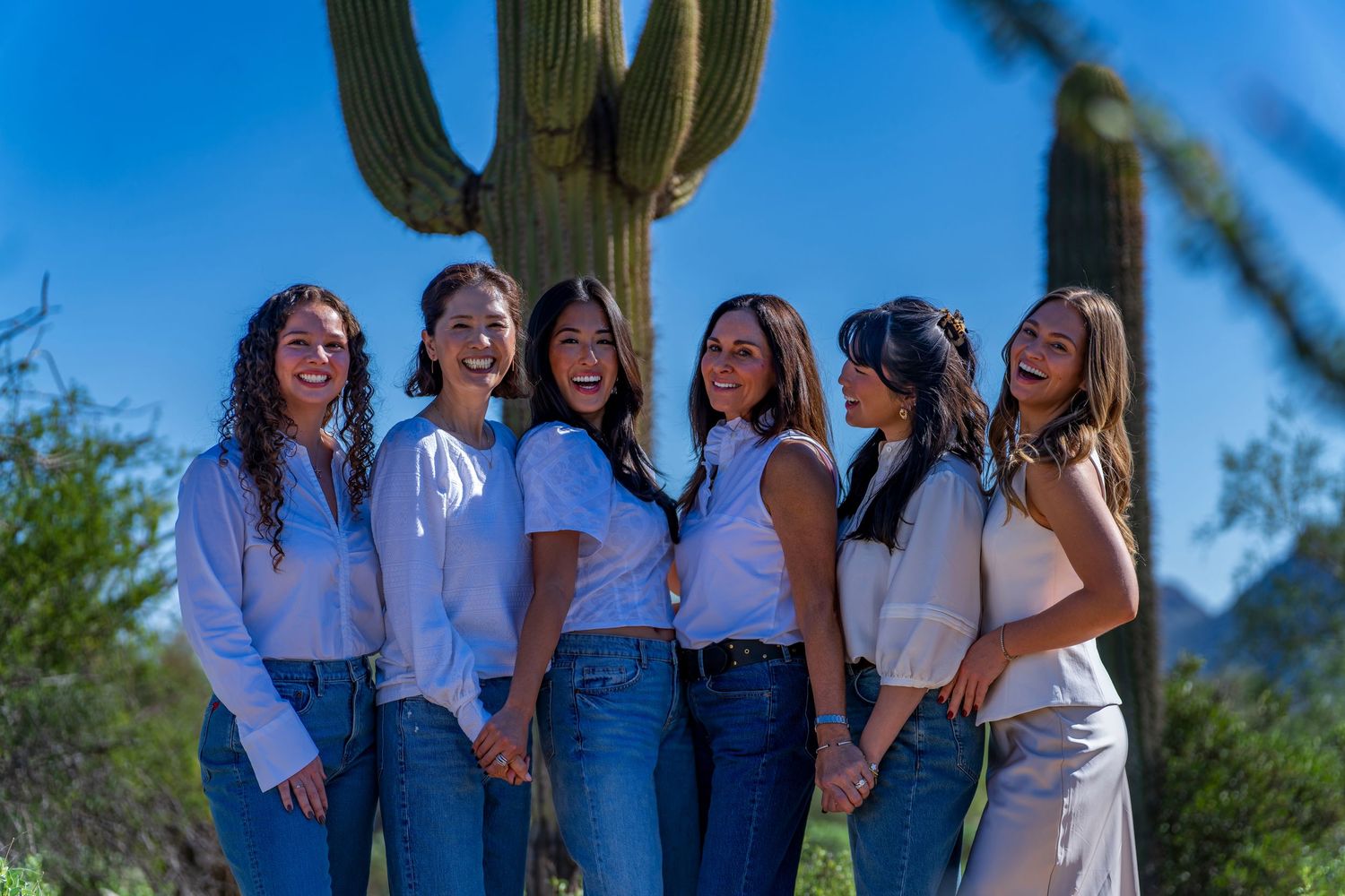 Women in coordinated white tops and denim posing by desert cactus.