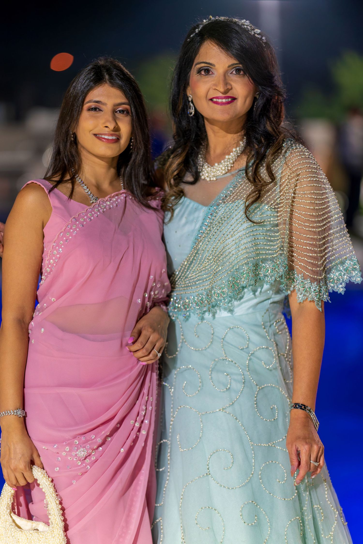Two women in elegant evening wear, one in pink sari and one in mint gown, pose together at formal event.