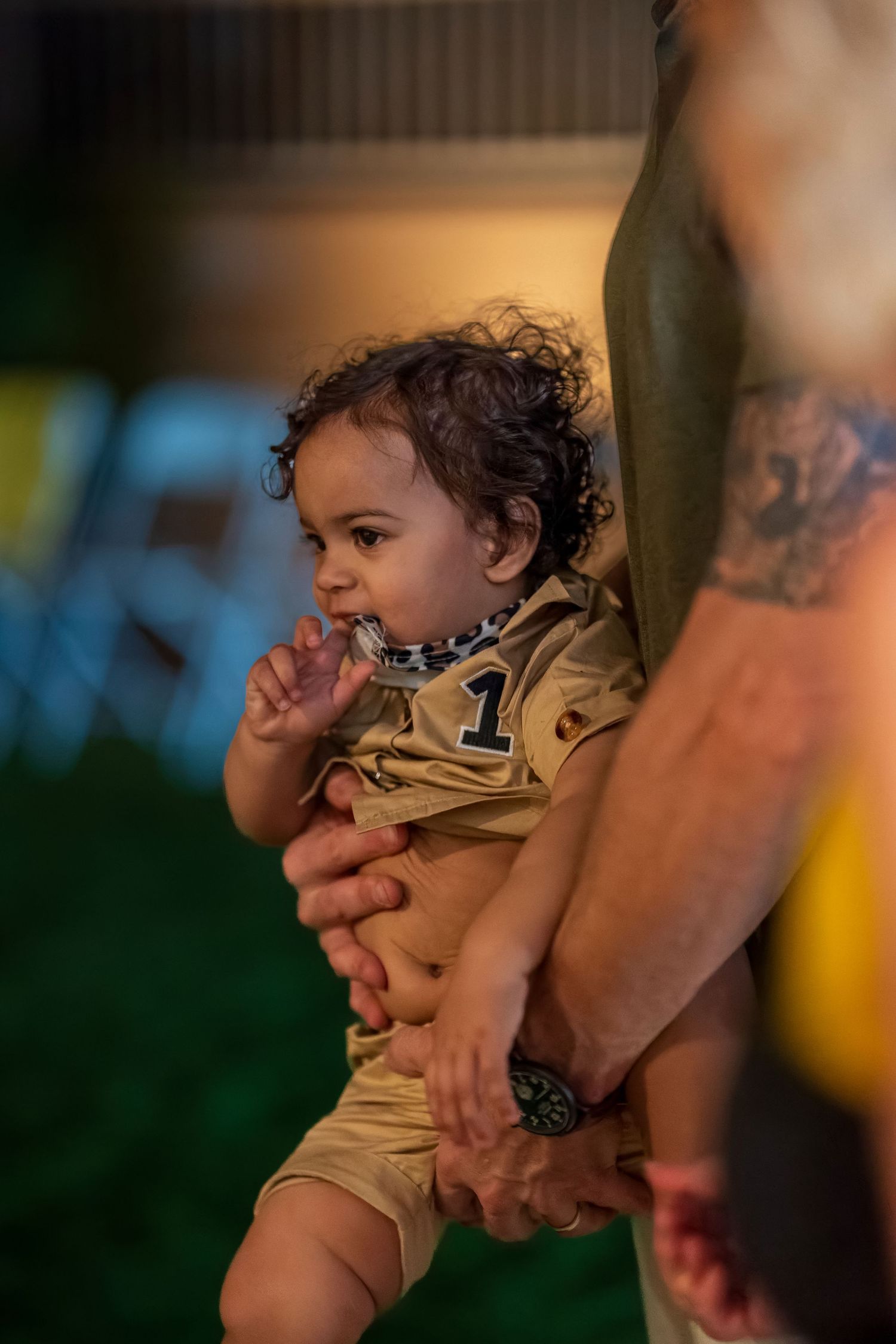 A curly-haired toddler in a beige outfit being supported while standing.