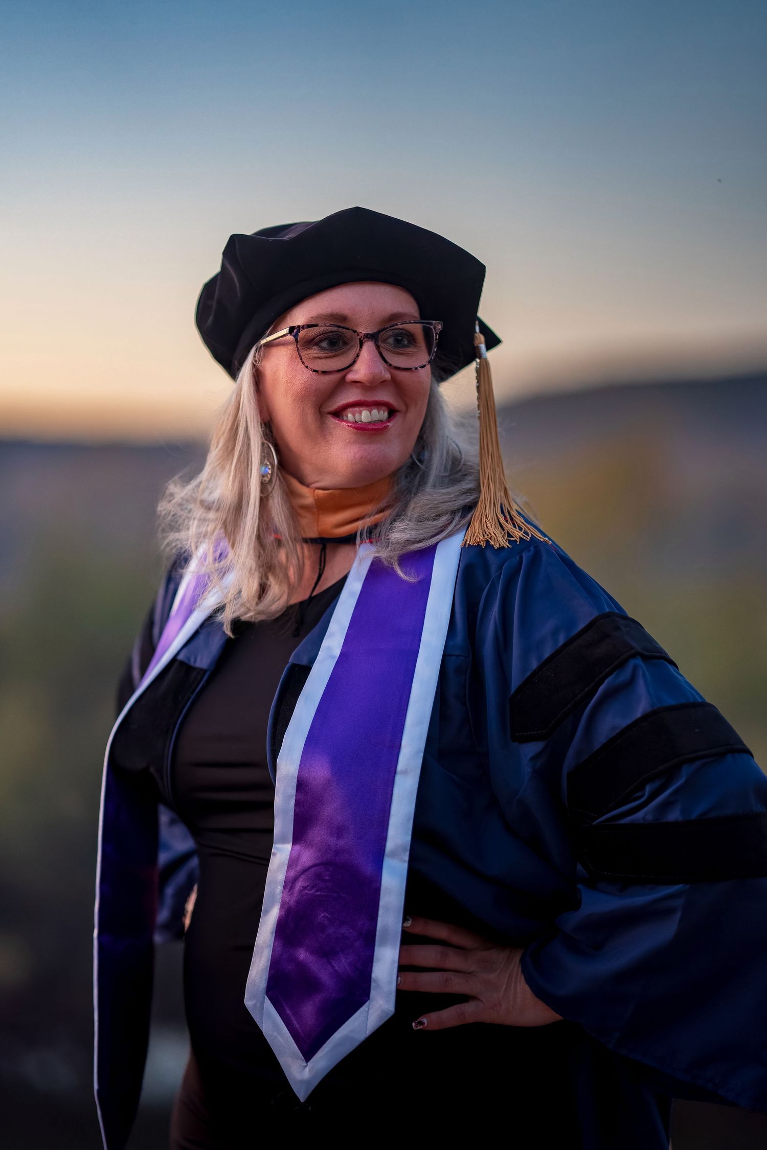 Graduate in doctoral regalia poses at sunset with mountains in background.