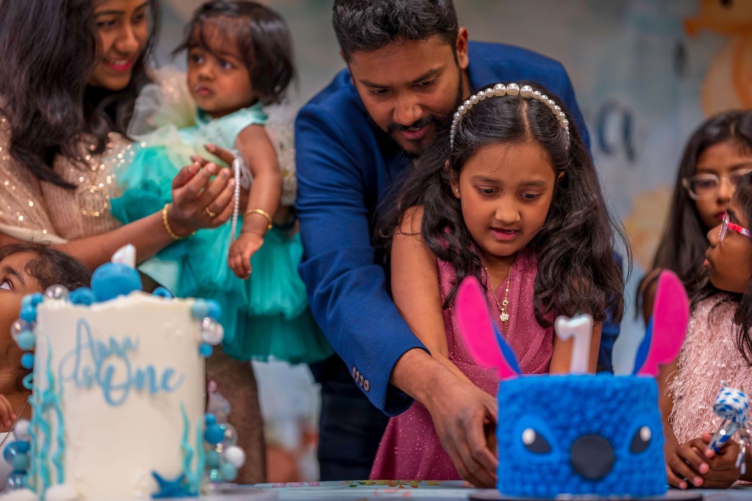 Family celebrating a first birthday party, cutting a decorated cake with a blue bunny theme.