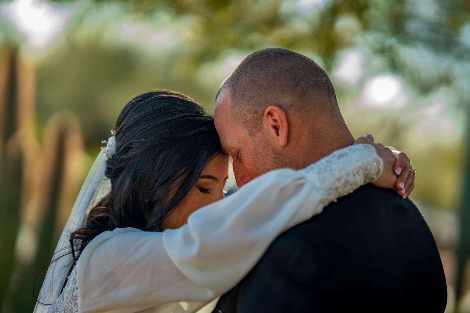 Emotional bride and groom embrace tenderly, foreheads touching, surrounded by lush green desert foliage.