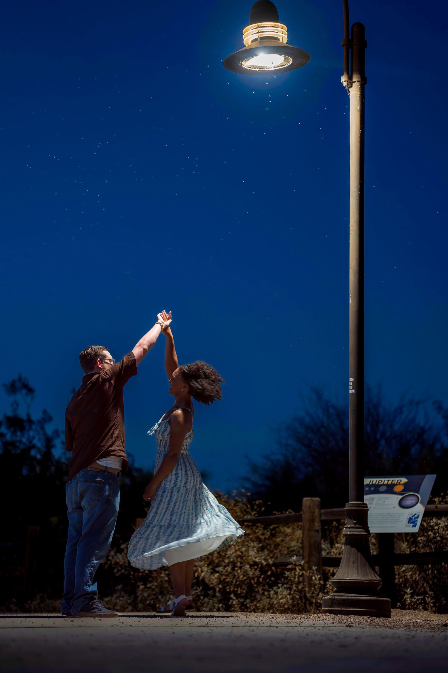 Couple dancing under a glowing street lamp at night, reaching up toward the light in a romantic outdoor setting.