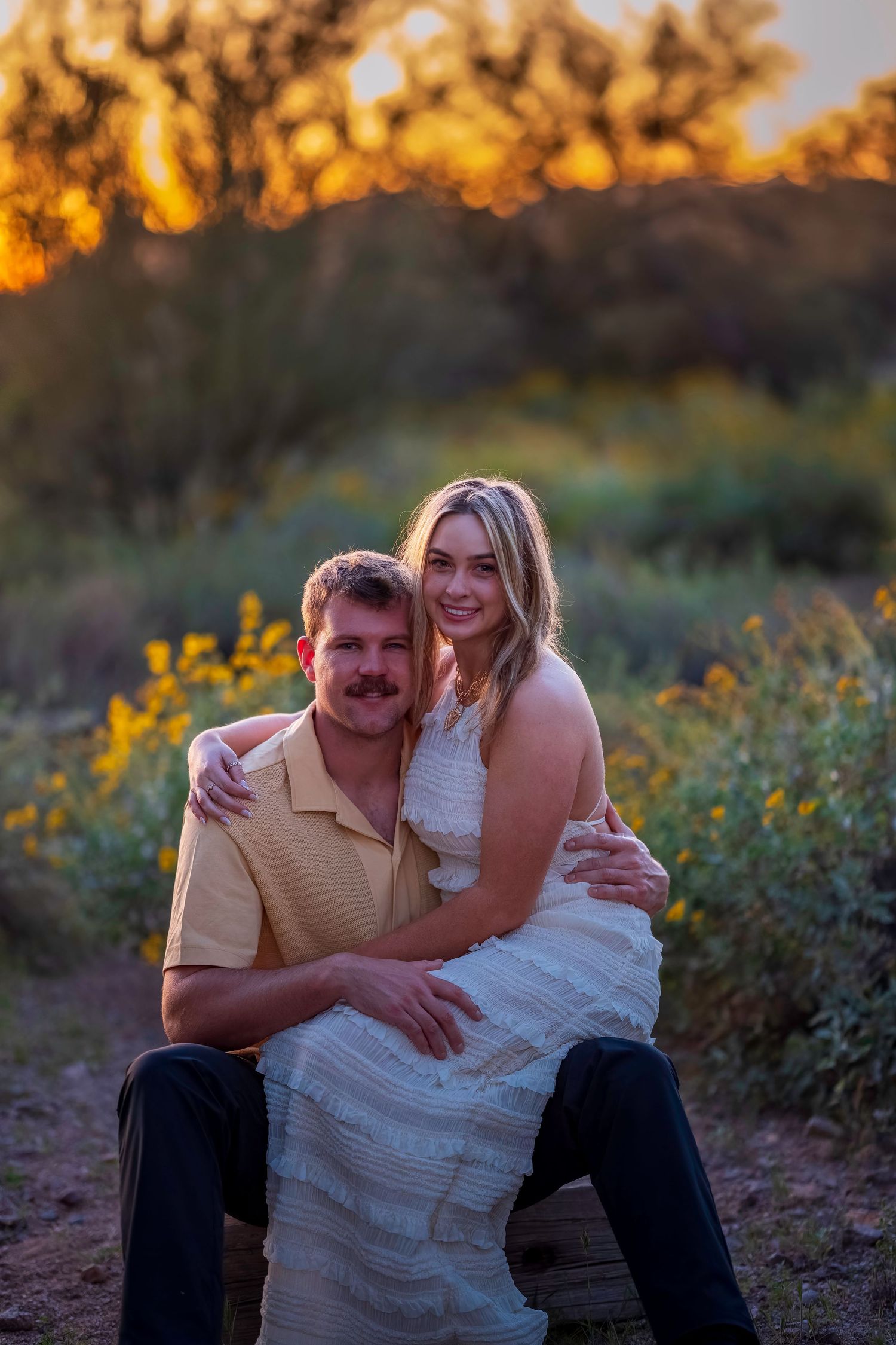 Couple posing at golden hour sunset in a field of yellow wildflowers, woman in white dress embracing man in yellow polo.