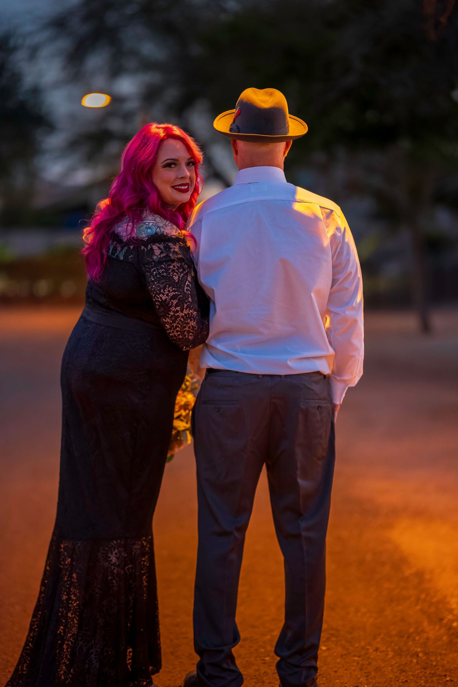 Couple standing together at night, woman with pink hair in black lace dress and man in white shirt and fedora.
