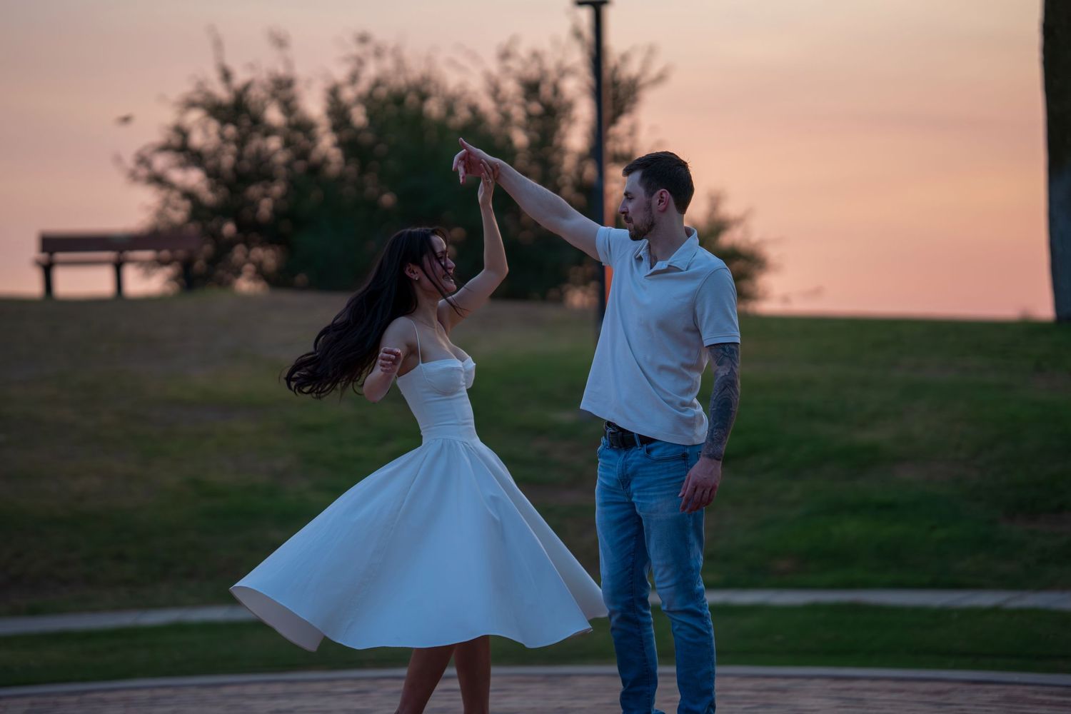 Couple dancing joyfully at sunset in a park, woman spinning in a white dress.